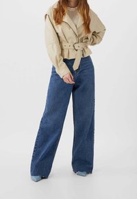 Beige cropped jas met een riem, gecombineerd met wijde blauwe denim jeans. De jas heeft een ritsluiting en schouderdetails.