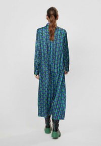 Lång, avslappnad klänning i mörkblått med ett geometriskt mönster i grönt. Har långa ärmar och krage, kombinerad med svarta platåstövlar.