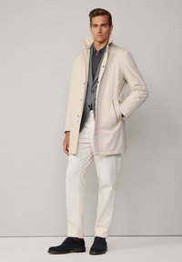 Chaqueta impermeable de color beige claro con un cuello alto, combinada con una camisa oscura, pantalones blancos y zapatos de cordones oscuros. Textura suave, diseño minimalista.