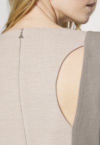 Rochie maro deschis, cu o decupare mare la umărul stâng, dintr-un material texturat și dotată cu accesorii delicate din argint la gâtul din spate.