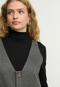 Chaleco de punto gris con un diseño de escote en V, adornado con un botón central, llevado sobre un suéter de cuello vuelto negro de canalé. Textura de tela suave.
