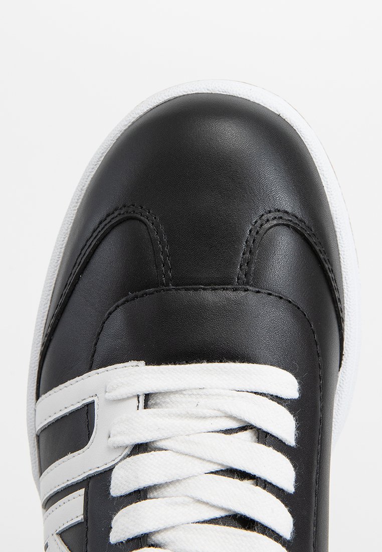 Zapatilla de cuero negro con punta redondeada, cordones blancos y suela de goma blanca; presenta costuras en contraste y detalles de marca.