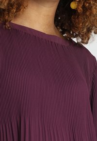 Närbild på en person som bär en plisserad, mörklila topp med rund halsringning och gula runda örhängen, med lockiga hårstrån synliga.