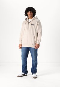 Beige zip-up hoodie met een grote voorzak, zachte textuur en zwart logo detail. Draag het over een wit shirt, in combinatie met blauwe spijkerbroeken en sneakers.