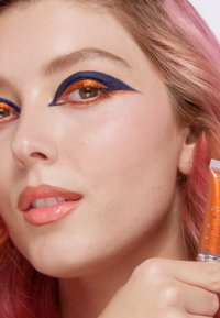 Tube de cosmétique tenu près du visage d'un modèle ; présente un maquillage des yeux audacieux en bleu marine et orange, des lèvres pêches et des cheveux rose doux. Texture de peau lisse.
