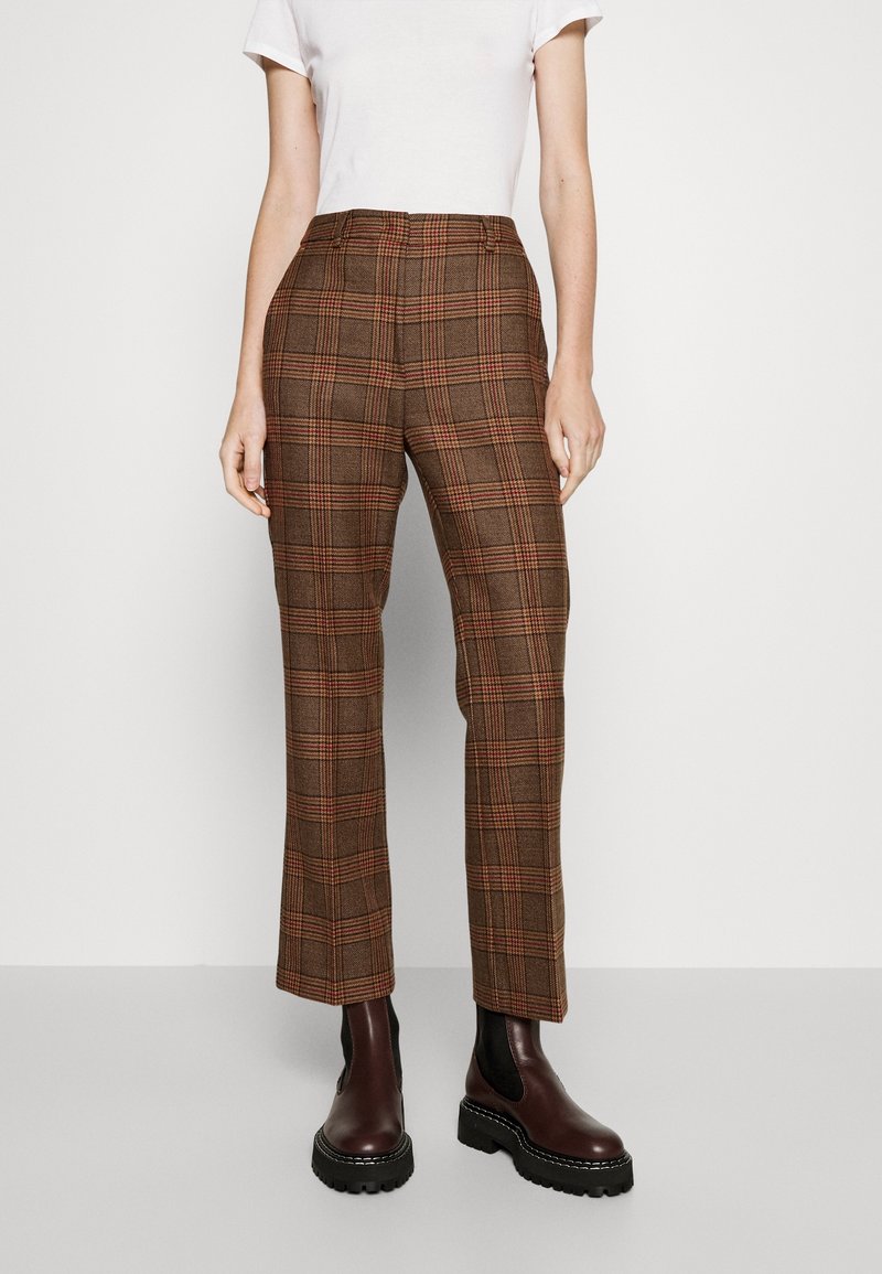 Femme portant un pantalon droit à carreaux marron et des bottes épaisses marron foncé, debout devant un fond clair uni.