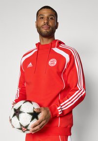 Rød atletisk hættetrøje med hvide detaljer og tre hvide striber på ærmerne, med FC Bayern München logo; holder en fodbold med sorte stjerner.
