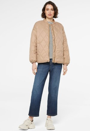 Frau in beige gesteppter Jacke über einem schwarz-weiß gestreiften Hemd, blauen Jeans und weißen Turnschuhen, die vor einem weißen Hintergrund steht.