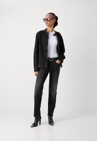 Giacca in denim nera, top bianco a coste, jeans neri a vita alta e stivali neri alla caviglia con punta affusolata. Modella che indossa occhiali da sole grandi.