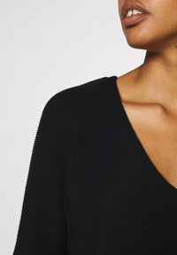 Pull en maille noire avec un décolleté en V profond, un tissu texturé et des manches raglan, mettant en valeur une partie de la clavicule et de l'épaule supérieure.