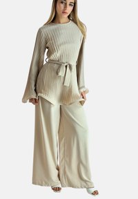Conjunto de tela mixta beige con pliegues verticales, cintura ceñida con cinturón, mangas largas, pantalones de pierna ancha y tacones abiertos blancos. Textura suave.
