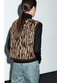Brun och krämrandig utan ärmar väst gjord av ull, med en fluffig textur. Bärs över en svart långärmad topp med blå denimbyxor.