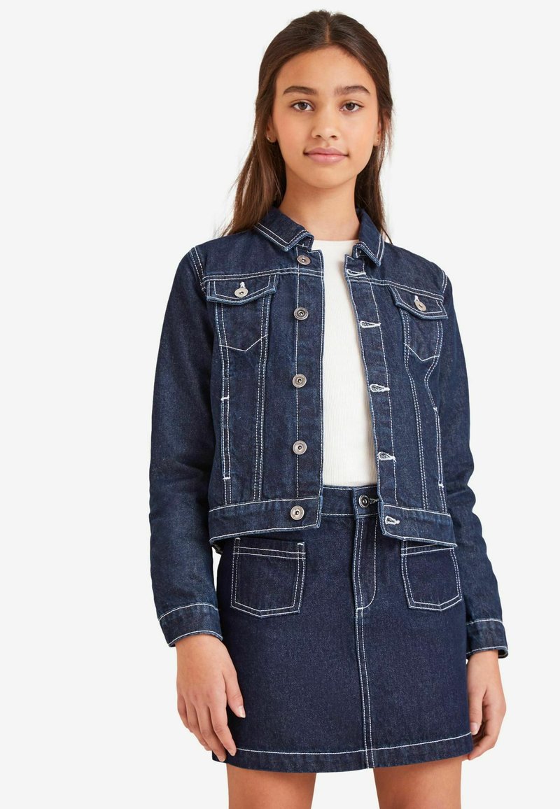 Temno moder jeans jakna z kontrastnimi belimi šivi, dvema sprednjima žepoma in srebrnimi gumbi, v kombinaciji z usklajenim jeans krilom.