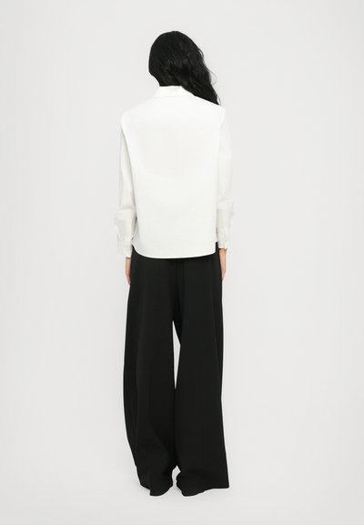 Cămașă albă cu guler, cu mâneci lungi, asortată cu pantaloni negri, largi și fluizi, evidențiind un design minimalist și o textură de țesătură fină.