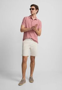 Camisa de polo rosa claro, pantalones cortos crema, zapatos grises. El modelo lleva gafas de sol y tiene el cabello castaño claro. Estilo casual contra un fondo neutral.