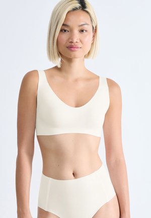 Femme aux cheveux blonds courts portant un soutien-gorge de sport crème sans coutures et une culotte taille haute sur un fond clair.