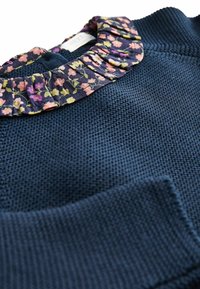 Pull en maille bleu marine avec un col froufroutant à motif floral dans des nuances de rose, vert et violet, avec une petite étiquette de marque blanche.