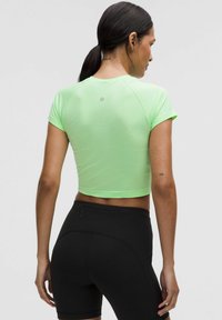 Neonově zelený sportovní crop top se krátkými rukávy a texturovanou tkaninou, zkombinovaný s černými těsnými šortkami, které mají diskrétní kapsu na zip na zadní straně.