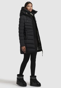 Zwarte pufferjas met een hoge kraag, ritssluiting en zijzakken. Gecombineerd met zwarte leggings en chunky zwarte laarzen, tegen een effen achtergrond.
