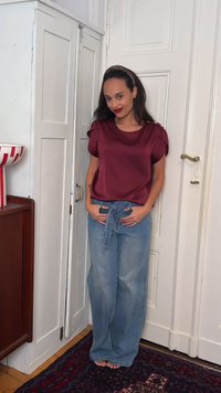 Γυναίκα με μακριά σκοτεινά μαλλιά και κορδέλα με λεοπάρ σχέδιο, φορώντας μπορντό σατέν τοπ και φαρδιά τζιν με δέσιμο στη μέση, στέκεται ξυπόλητη μέσα στο σπίτι.