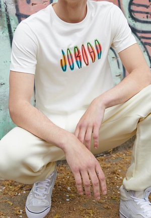 Junge Person in Hockstellung, trägt weiße Hose, weiße Turnschuhe und ein weißes T-Shirt mit buntem "JORDAN"-Schriftzug vor einer Graffiti-Wand.