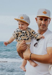 Casquette grise avec des accents jaunes, pull beige avec des imprimés de panda noirs, shorts beige clair ; adulte tenant un enfant sur la plage avec une vue sur l'océan.