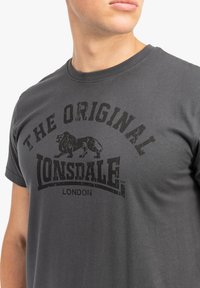 Szürke pamut póló, kerek nyakkal, melyen egy nagy fekete grafikai nyomat található, a "THE ORIGINAL LONSDALE" felirattal és egy oroszlánmintával.