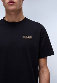 Black cotton t-shirt featuring a rectangular embroidered patch with "NAPAPIJRI" and "geographic" in contrasting colors on the left chest.