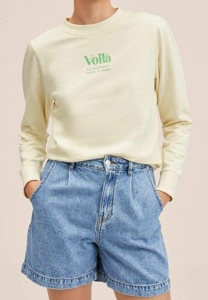 Personne portant un t-shirt crème à manches longues avec un texte vert et un short en jean bleu ample taille haute, les mains dans les poches.