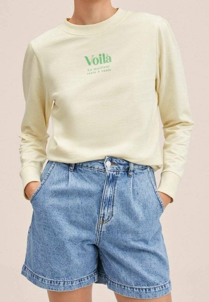 Personne portant un t-shirt crème à manches longues avec un texte vert et un short en jean bleu ample taille haute, les mains dans les poches.