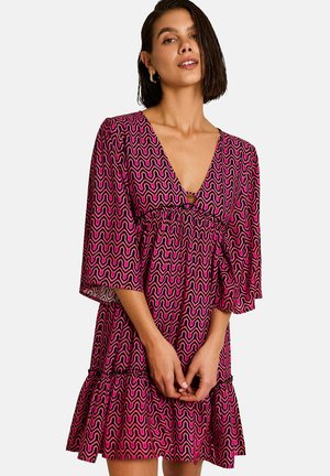 Femme aux cheveux courts et foncés portant une robe rose et noire à motif ondulé, manches trois-quarts et profond décolleté en V.
