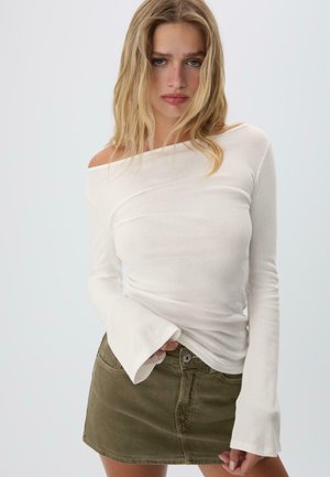 Donna bionda che indossa una maglietta bianca con le spalle scoperte e maniche lunghe, insieme a una mini gonna verde oliva, in posa davanti a uno sfondo chiaro e uniforme.