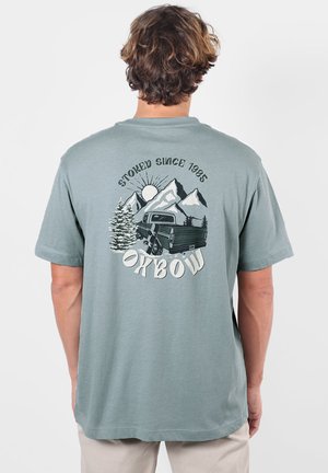 Homme portant un T-shirt vert clair-gris avec un graphique de montagnes, soleil, pins et pick-up, avec le texte "Stoked Since 1985 Oxbow" au dos.