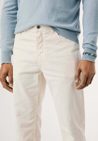 Pantaloni in corduroy chiaro con texture verticale, abbinati a un maglione blu, con polsini a coste e un dettaglio logo sulla manica.