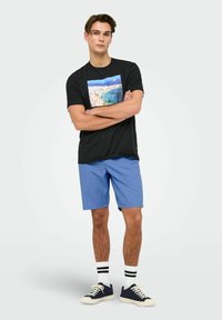 T-shirt nera con stampa di una scena da spiaggia, abbinata a pantaloni blu. L'outfit presenta calze bianche con strisce nere e scarpe sportive blu navy.