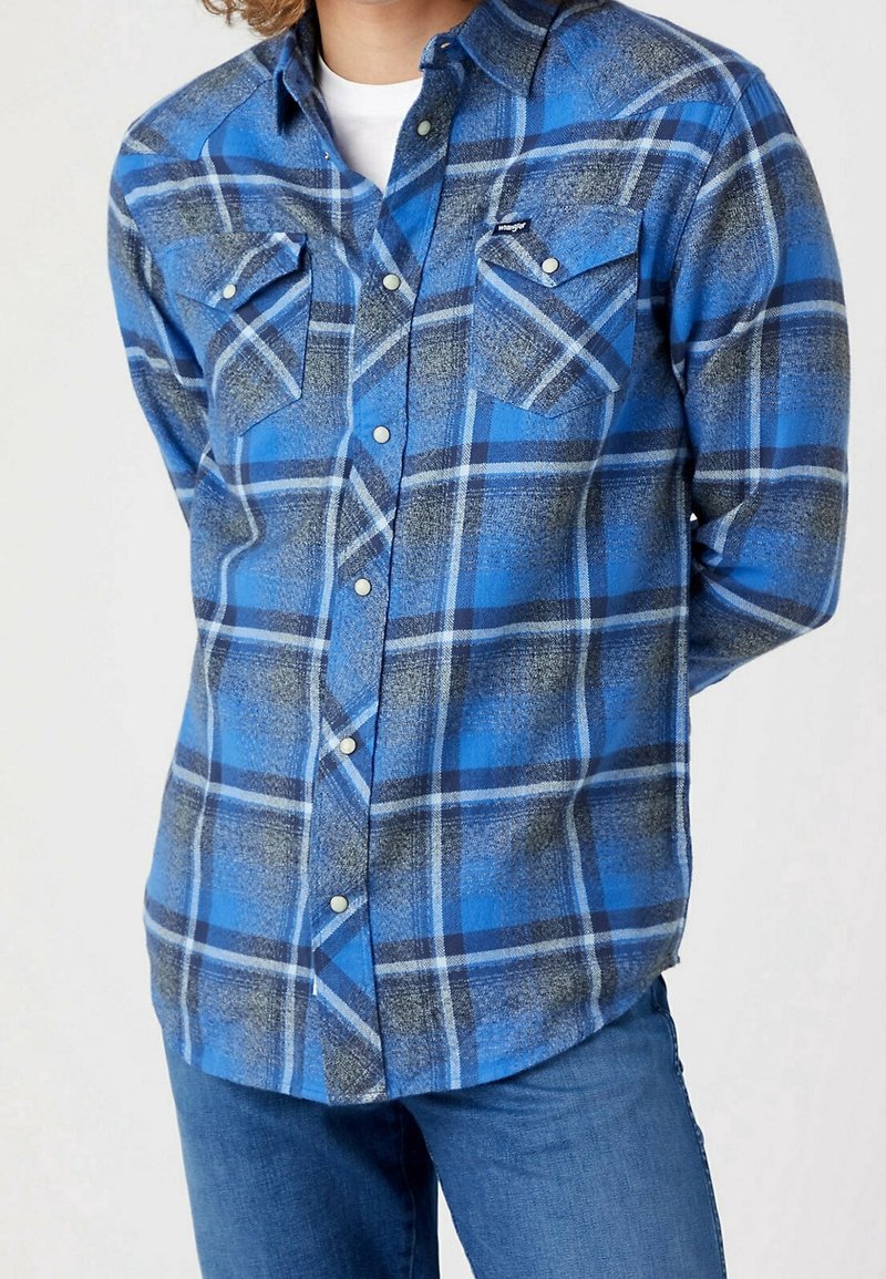 Personne portant une chemise à carreaux bleue, grise et blanche boutonnée sur un sous-maillot blanc avec un jean bleu, debout les mains dans le dos.