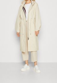 Giacca beige con cappuccio, cordino in vita, maniche lunghe e zip frontale. Abbinata a pantaloni chiari e sneakers bianche. Texture liscia.