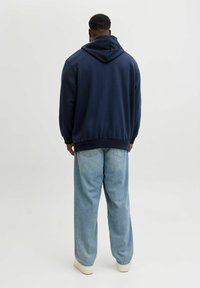 Navyblauwe hoodie met een grote capuchon, gestructureerde stof en ribgebreide zoom, gecombineerd met lichtblauwe wijde spijkerbroek en witte sneakers.