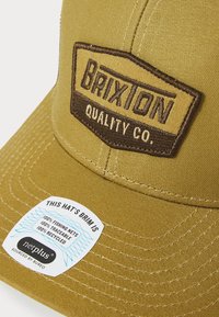 Brown and khaki baseball cap featuring a stitched logo patch and a label indicating the brim is made from recycled fishing nets.