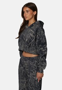 Krótkie bluza z kapturem w szarym i czarnym wzorze moro, uszyta z miękkiego materiału. Posiada frontową kieszeń i kaptur ze sznurkiem. Połączona z dopasowanymi dresami.