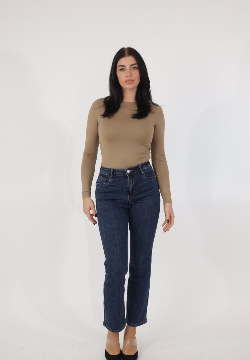 Mujer con cabello largo y oscuro, vestida con una camiseta beige de manga larga, jeans azules y tacones negros, de pie frente a un fondo blanco liso.