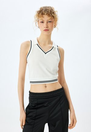 Top corto sin mangas de polo blanco con escote en V y detalles de rayas negras en el cuello y el dobladillo, combinado con pantalones deportivos negros.
