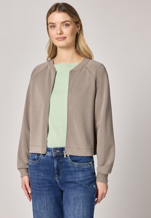 Mladá žena v taupe bundě na zip přes světle zelené tričko a modré džíny na jednobarevném světle šedém pozadí.