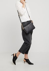 Borsa a tracolla in pelle nera con dettagli in catena dorata. Forma rettangolare, texture liscia. Indossata con una camicetta bianca leggera e jeans grigi.