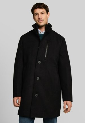 Uomo che indossa un cappotto nero con bottoni, tasca sul petto con cerniera e collo alto, in piedi su uno sfondo grigio uniforme.