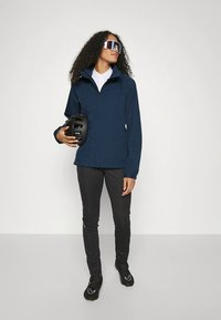 Tamsiai mėlyna neperšlampama striukė su aukštu apykakle, derinama su aptemptomis juodomis kelnėmis ir juodu šalmu. Išvaizdą papildo didelės kvadratinės saulės akiniai.