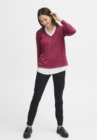 Maglione bordeaux a scollo a V sovrapposto a una camicia bianca, abbinato a pantaloni slim neri e mocassini neri, su uno sfondo neutro.