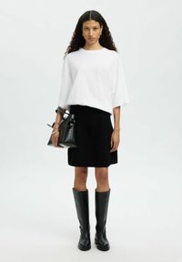Weißes, übergroßes T-Shirt, schwarzer knielanger Rock, schwarze hohe Stiefel, hält eine schwarze Ledertasche mit minimalistischem Design und Riemen.