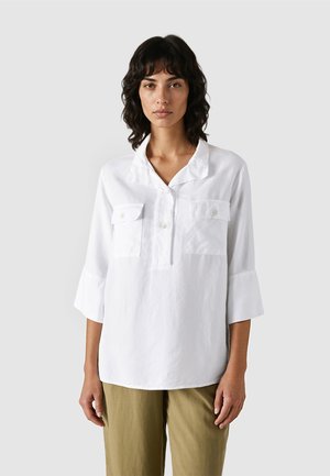 Frau mit mittellangem, dunklem, welligem Haar trägt ein weißes, langärmliges Hemd mit Knöpfen und hellbraune Hose vor einem schlichten Hintergrund.