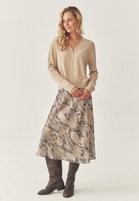 Kvinde med langt blond hår iført en beige lynlås cardigan, en midi-nederdel med slangeprint og mørkebrune knæhøje støvler, der står på en ensfarvet baggrund.